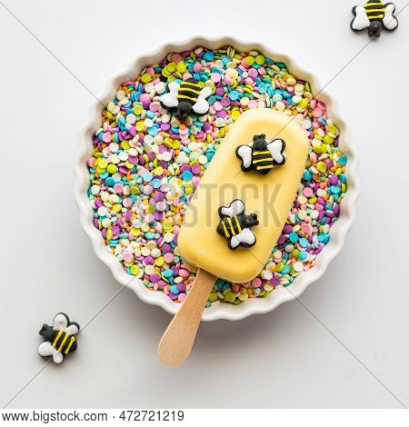 A Homemade Cakesicle Decorated With Edible Bees In A Bowl Full Of Sprinkles.