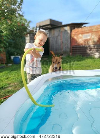 A Child Boy With A Dog Indulges In An Inflatable Pool Of Water On A Hot Sunny Summer Day. The Child 