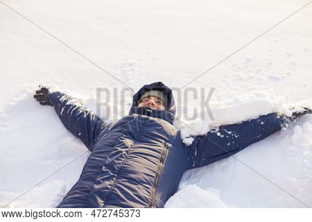 Male Man In Winter Clothes Lies Down In Soft White Snow. European Caucasian White Smiling Man Portra