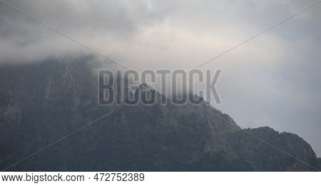 Dark Atmospheric Surreal Landscape With A Dark Rocky Mountain Peak In Low Clouds In A Gray Cloudy Sk