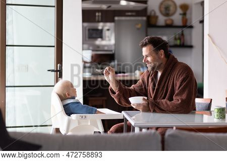 Father Wearing Bathrope Spoon Feeding Hir Infant Baby Boy Child Sitting In High Chair At The Dining 