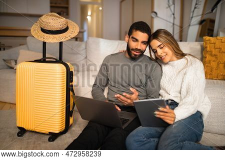 Smiling Multiracial Young Couple Sit On Floor With Gadgets, Suitcase And Hat, Choose Country For Vac