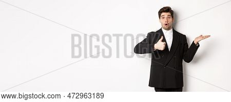 Portrait Of Hadnsome Bearded Man In Formal Suit, Showing Thumb-up And Holding Product In Hand Over W