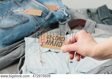 Clothes From Females Wardrobe On Shelf Of Second Hand Store. Womans Hand Holding Craft Paper Tag Wit