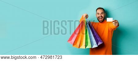 Happy Aduly Man Showing Credit Card And Shopping Bags, Standing Against Turquoise Background