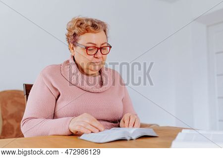 A Senior Woman, Glasses On Her Face, Reads A Book In Her Cozy Living Room, The Pages Turning Steadil