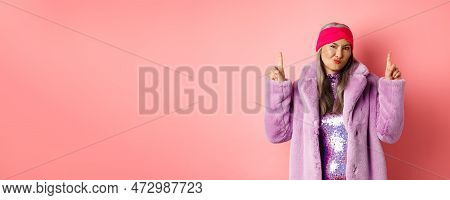 Disappointed Stylish Asian Lady Pointing Fingers Up. Senior Woman In Trendy Fake Fur Coat Grimacing 