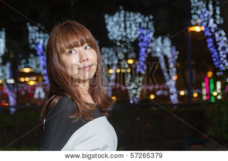 Asian Woman Standing In A Park With Lights Behind Her