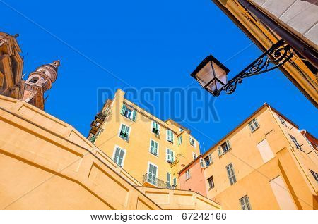 Yellow and orange residential houses and bell tower of Saint Michel Archange basilica under clear blue sky in Menton, France.