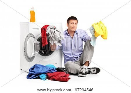 Confused young man doing laundry seated by a washing machine isolated on white background