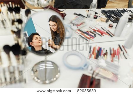 professional makeup artist doing anti-aging makeup for middle aged women. Make-up artist work in beauty salon. Real people. Reflection in the mirror as visagiste applying makeup. Backstage photo