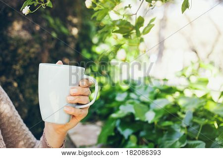 Close-up of woman hand with cup of coffee. Early morning routine. Green garden on the background. Selective focus film effect .