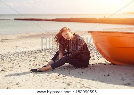 A girl sideways sitting near a red boat on the beach by the sea with beautiful sunset on the background. Woman in jeans pullover and jacket on the sand
