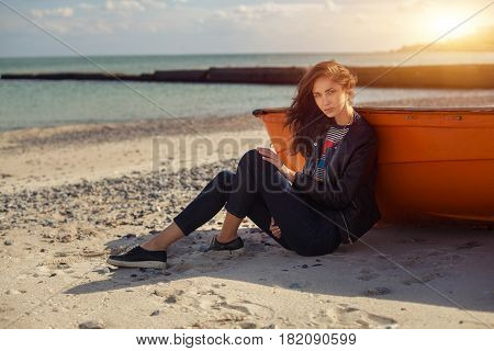 A girl sideways sitting near a red boat on the beach by the sea with beautiful sunset on the background. Woman in jeans pullover and jacket on the sand