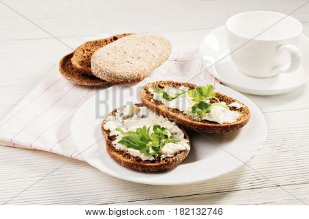 Rye bread with soft cheese and greens. Composition in rural style white ware on a white wooden table. The food lies on a napkin from fabric. Selective focus