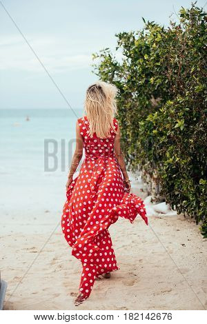 Beautiful young woman with long blond hair in long red dress goes along the beach. Girl in red dress. Long red dress flutters in the wind