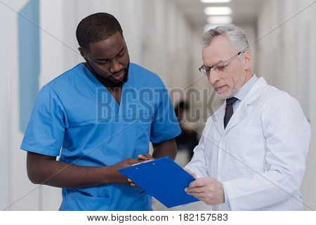 Teaching my intern. Aging professional positive doctor standing in the hospital while holding folder and consulting African American intern