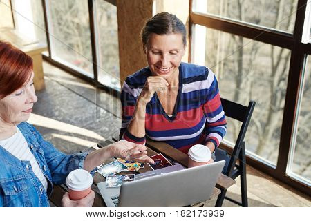 Female pensioner pointing at laptop display while showing friend images of her relatives