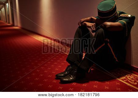 Tired hotel servant sitting by wall in hotel corridor