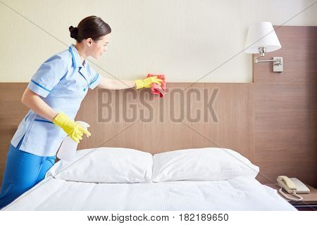 Pretty chambermaid dusting bed with sponge in hotel-room