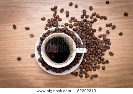 White cup with black, hot coffee standing on brown, roasted beans on wood table, close up.