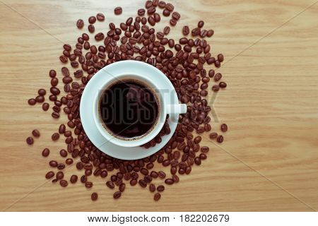 White cup with black, hot coffee standing on brown, roasted beans on wood table, close up
