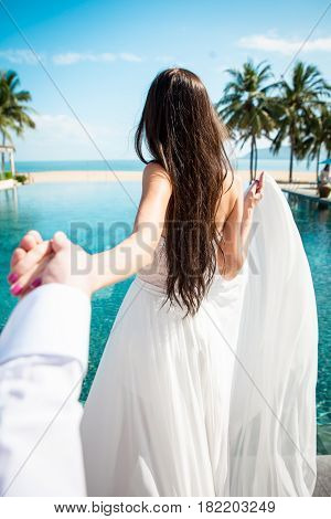 Sexy bride in white dress holding groom in luxury resort. Romantic woman leading man to swimming pool. Fiance follow fiancee. Tropical country. Caucasian model with long brown hair.