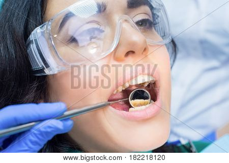 Stomatologist is using concave mirror. Female patient in safety glasses. Dental examination procedure.