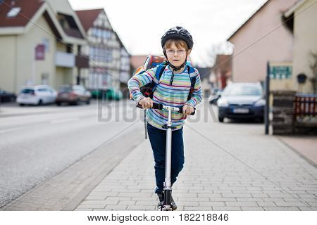 Active school kid boy in safety helmet riding with his scooter in the city with backpack on sunny day. Happy child in colorful clothes biking on way to school. Safe way for kids outdoors to school.