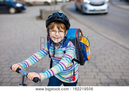 Active school kid boy in safety helmet riding with his scooter in the city with backpack on sunny day. Happy child in colorful clothes biking on way to school. Safe way for kids outdoors to school.