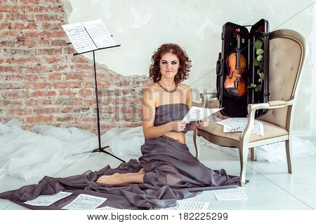 Beautiful woman sitting on the floor near the chair with violin in case and holding notes in hand
