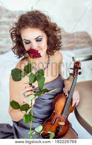 Front view close up beautiful woman sitting on the floor near chair and holding a violin and smelling red rose flower in studio