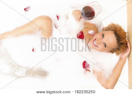 Spa Concepts. Sensual Alluring Sexy Caucasian Blond Female in Foamy Bathtub Filled with Flowery Petals Relaxing Holding a Glass of Red Wine. Passionate Look. Horizontal Image
