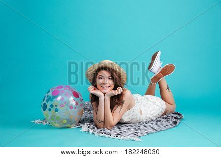 Portrait of a cheerful smiling summer girl in straw hat laying on a blanket with beachball isolated over blue background