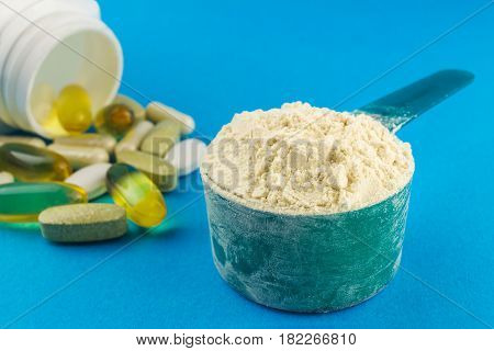 Protein powder in measuring plastic spoon and vitamin complex omega 3 glucosamine capsules supplements on blue background sports nutrition.