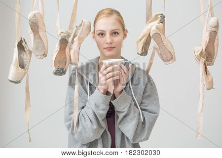 Portrait of a young ballerina with a cup of tea in her hands is among the hanging ballet slippers. Pointe shoes are arranged in random order.
