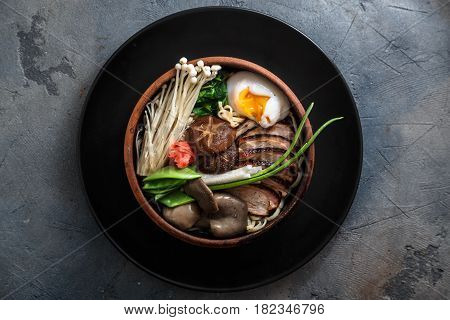Ramen noodles with duck, egg, enoki and shiitake mushrooms with broth on dark background, top view