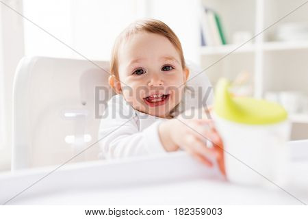 childhood and people concept - happy smiling little baby drinking from spout cup sitting in highchair at home