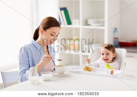 family, eating, breakfast and people concept - happy mother adding sugar to coffee cup and little baby with food sitting in highchair at home kitchen