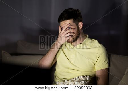 people, mass media, television and entertainment concept - scared young man watching tv and eating popcorn at night at home