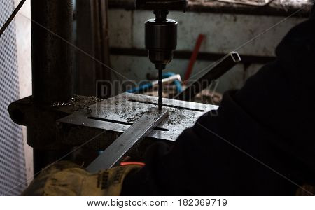 Construction worker drills hole in rod of metal iron using industrial drill in a dark dingy workshop / workshed / factory