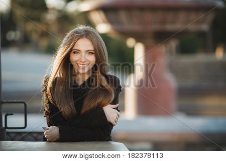 Young,beautiful,attractive woman,brunette with long straight hair and gray eyes,a beautiful smile and straight white teeth,dressed in a black knitted pullover, sitting at a table in the street cafe,posing against a background of an urban landscape.