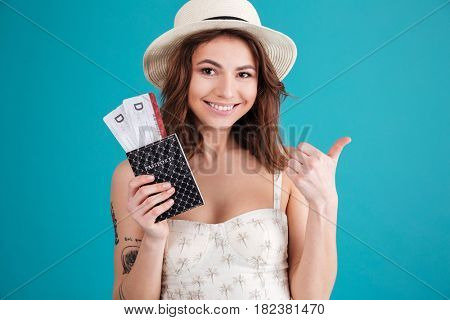 Close up portrait of a smiling happy woman traveller in summer clothes holding passport with plane tickets and showing thumbs up gesture isolated over blue background