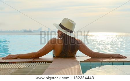 Woman in big hat relaxing on the swimming pool near the sea in the sunset