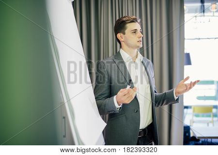 Confident Young Team Leader Or Manager Standing In Front Of A Flip Chart And Present Project On Offi