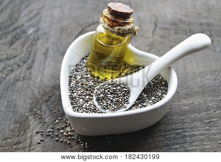 Chia seeds and chia oil in a white heart shaped bowl on old wooden background. Organic chia seed oil. Salvia hispanica seeds. Healthy food, superfood or bodycare concept. Selective focus.