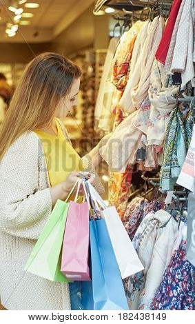 Vertical shot of woman holding shopping bags ino one hand looking at shawls hanging inside a cloth store.