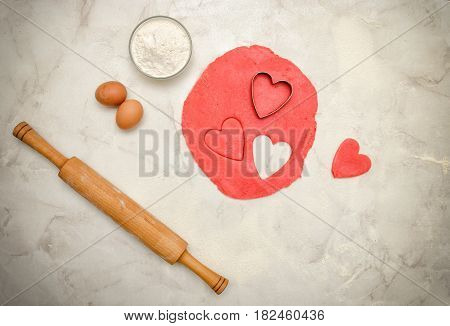 Red dough with a cut out hearts eggs flour and rolling pinon a white table. Top view