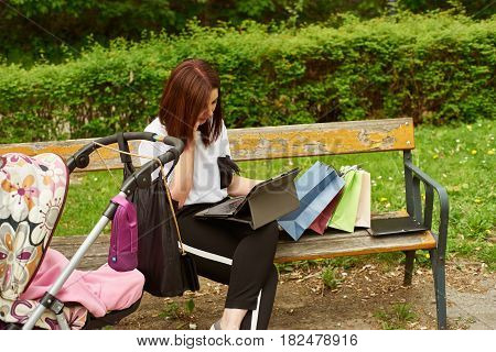 A woman with baby carriage and shopping bags is speaking on the phone and working with tablett in the park, while sitting on a bench.