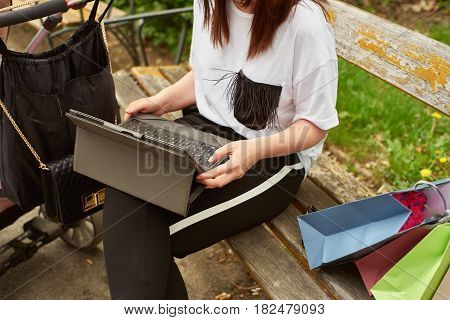 Crop shot of a woman with baby carriage and shopping bags working with tablett in the park while sitting on a bench.